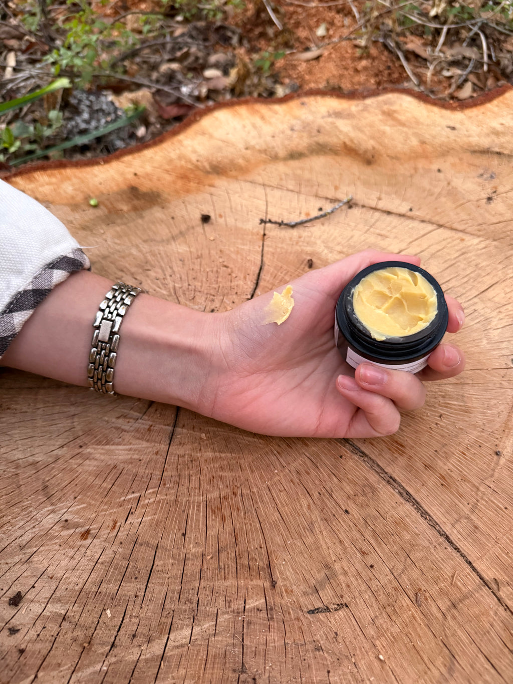 A hand with a silver link bracelet and plaid shirt cuff holds a black jar of rich yellow cream, with a small swatch on the skin, resting on a cracked tree stump with visible growth rings and blurry natural foliage