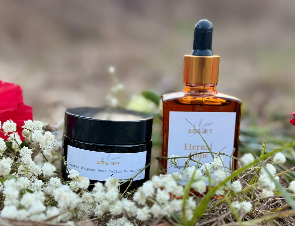 SOS 4:7 skincare products including an amber Eternal Glow dropper bottle with a gold cap and a black Organic Whipped Beef Tallow Moisturizer jar presented outdoors amongst white baby's breath flowers and dry grass with a hint of red petals
