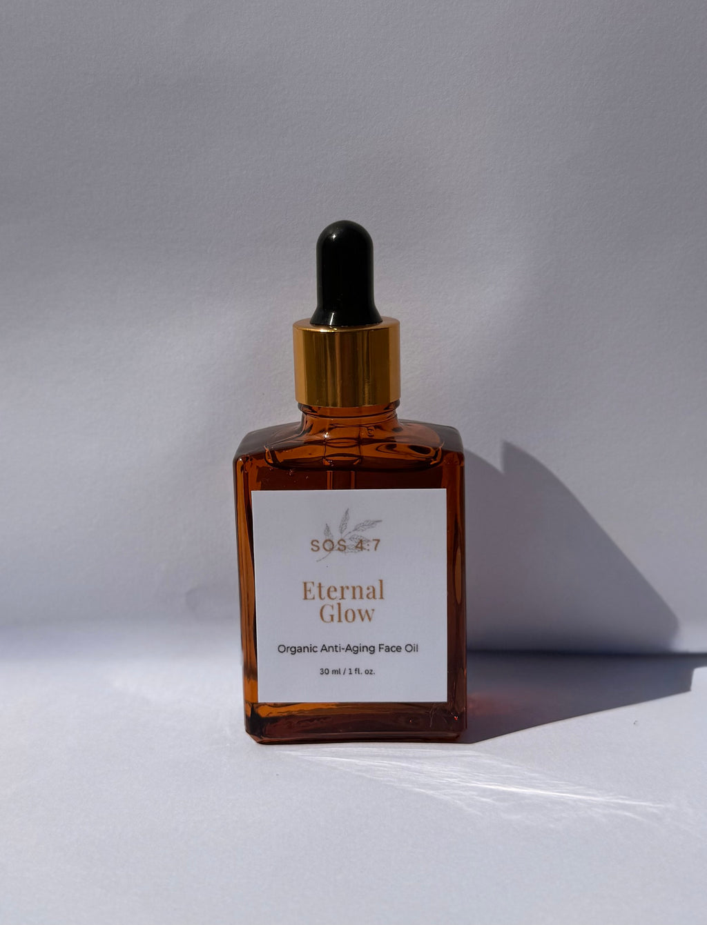 Photograph of an SOS 4:7 Eternal Glow Organic Anti-Aging Face Oil in an amber square glass bottle with a gold and black dropper, featuring a white label with a leaf motif, presented on a minimalist white background with a cast shadow