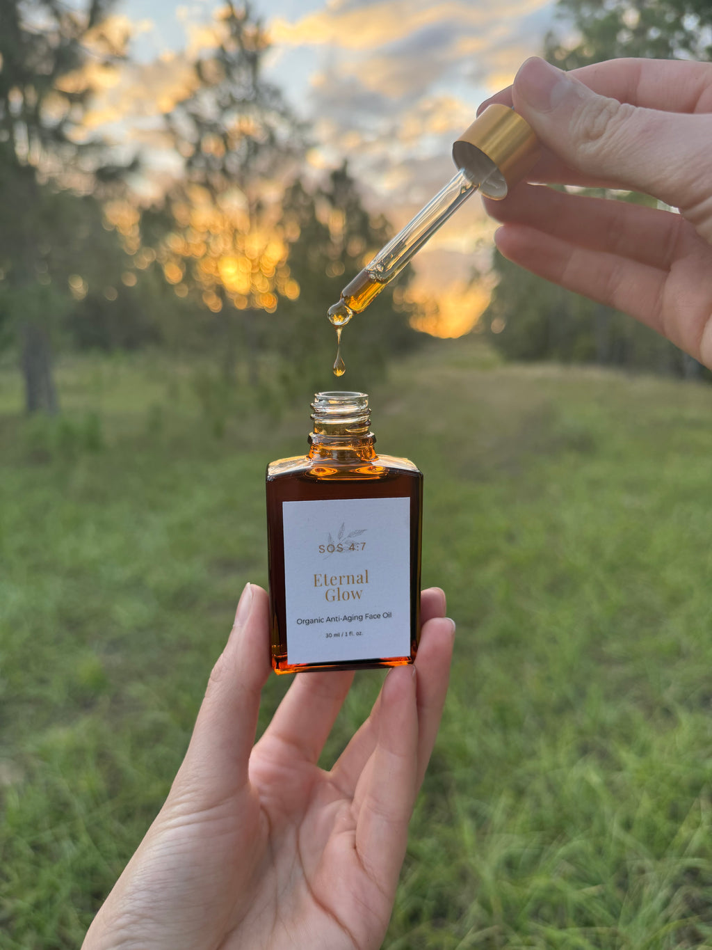 Photograph of hands holding an amber bottle of SOS 4:7 Eternal Glow Organic Anti-Aging Face Oil as a golden oil droplet falls from a dropper against a blurred warm sunset and green natural background