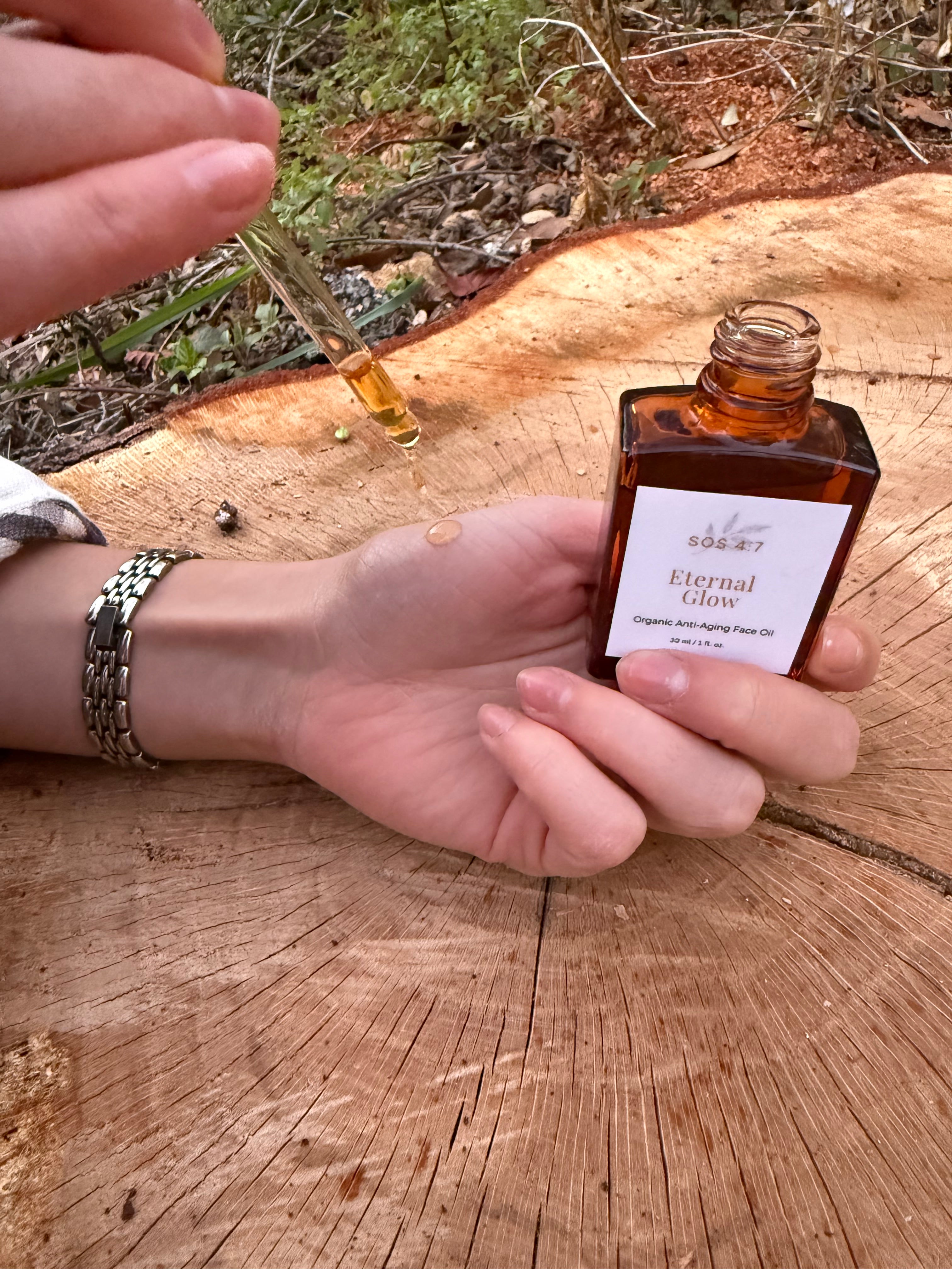 Photograph of a hand wearing a golden chain-link watch holding an amber SOS 47 Eternal Glow Organic Anti-Aging Face Oil bottle, dropping a golden oil droplet onto the palm over a rustic tree stump with green foliage