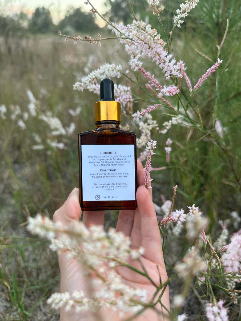 A hand holds a brown amber glass skincare bottle with a gold dropper and a white label displaying ingredients and SOS_47_SKIN brand details, nestled among delicate pink and white wildflowers in a natural outdoor setting