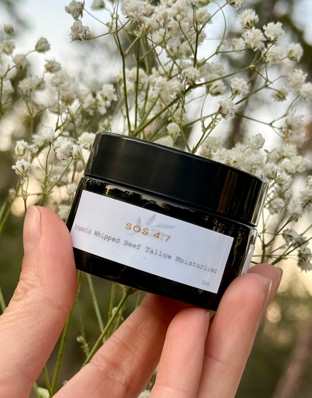 Photograph of a human hand holding a black jar of SOS 4:7 Organic Whipped Beef Tallow Moisturizer with a white label, set against a blurred background of delicate white baby's breath flowers