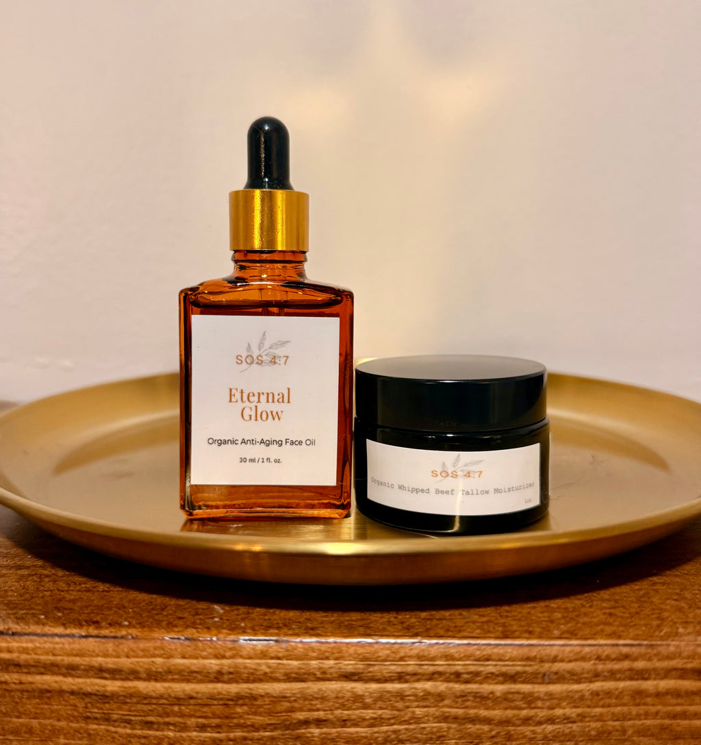 A still life photograph showcasing SOS 4:7 Eternal Glow Organic Anti-Aging Face Oil in an amber dropper bottle and Organic Whipped Beef Tallow Moisturizer in a black jar, both resting on a golden-brass circular tray on a wooden surface