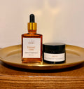 A still life photograph showcasing SOS 4:7 Eternal Glow Organic Anti-Aging Face Oil in an amber dropper bottle and Organic Whipped Beef Tallow Moisturizer in a black jar, both resting on a golden-brass circular tray on a wooden surface